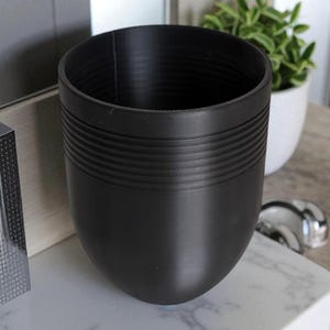 May include: A black, egg-shaped planter with horizontal ridged detailing. The planter is empty and sits on a white marble-patterned surface. A small potted plant is visible in the background.