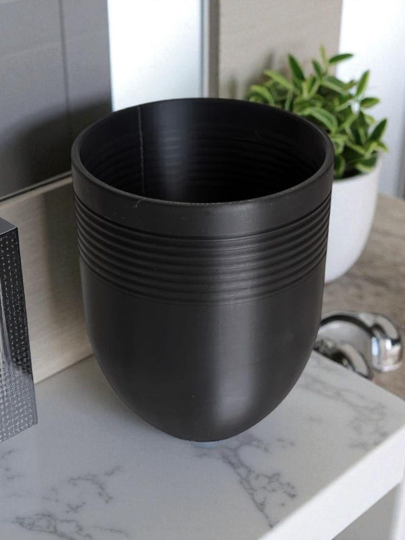 May include: A black, egg-shaped planter with horizontal ridged detailing. The planter is empty and sits on a white marble-patterned surface. A small potted plant is visible in the background.