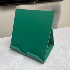 May include: A solid green book stand made of a single piece of material. The stand has a rectangular back and a triangular base, designed to hold books or tablets. The stand is on a light gray surface.