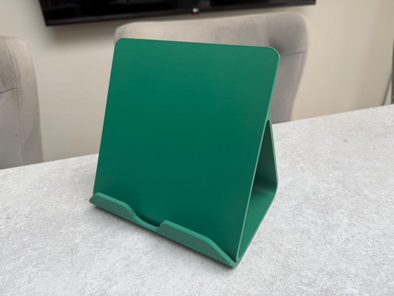 May include: A solid green book stand made of a single piece of material. The stand has a rectangular back and a triangular base, designed to hold books or tablets. The stand is on a light gray surface.