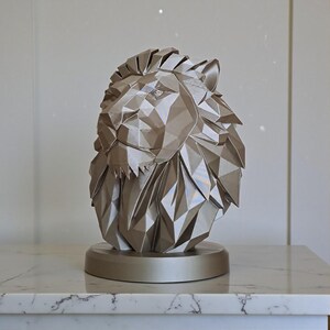 May include: A metallic gold lion head sculpture with a geometric, polygonal design. The lion's mane and facial features are intricately detailed. The sculpture is mounted on a circular base, creating a striking decorative piece.