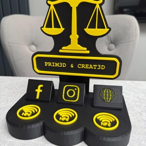 K&ouml;nnte beinhalten: Schwarz-gelber 3D-gedruckter Displayst&auml;nder mit einem Waagen-Design. Er verf&uuml;gt &uuml;ber Bereiche f&uuml;r Social-Media-Symbole und NFC-Symbole. Der Text "PRIM3D & CREAT3D" ist auf dem St&auml;nder zu sehen.
