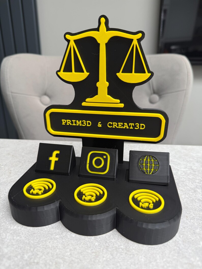 K&ouml;nnte beinhalten: Schwarz-gelber 3D-gedruckter Displayst&auml;nder mit einem Waagen-Design. Er verf&uuml;gt &uuml;ber Bereiche f&uuml;r Social-Media-Symbole und NFC-Symbole. Der Text "PRIM3D & CREAT3D" ist auf dem St&auml;nder zu sehen.