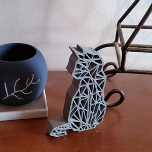 May include: A gray geometric cat sculpture with a detailed, open design. The cat is sitting, with its tail curled. A dark blue spherical vase with a white branch design is to the left. A brown metal decorative piece is in the background.