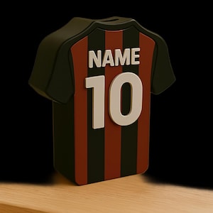 K&ouml;nnte beinhalten: Ein schwarz-rot-wei&szlig;es Sparschwein in Form eines Trikots. Das Trikot hat vertikale rote und schwarze Streifen, mit der Nummer 10 und dem Wort "NAME" in wei&szlig;en Buchstaben. Es steht auf einer Holzoberfl&auml;che.