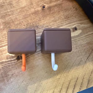 May include: Two brown square hooks with curved, colored ends. One hook has an orange end, and the other has a white end with a light blue tip. The hooks are on a wooden surface.