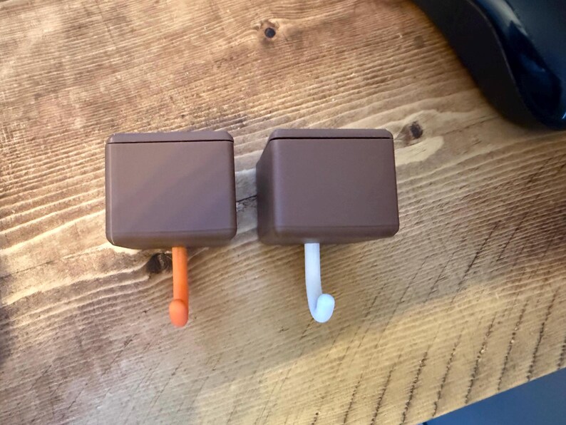 May include: Two brown square hooks with curved, colored ends. One hook has an orange end, and the other has a white end with a light blue tip. The hooks are on a wooden surface.