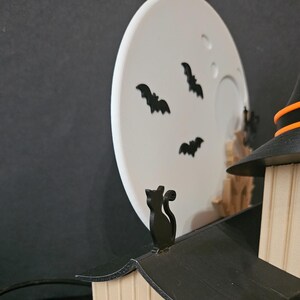 May include: A Halloween-themed decoration featuring a black cat on a rooftop, a witch's hat, and a white moon with black bat silhouettes. The scene is set against a dark background, creating a spooky ambiance.