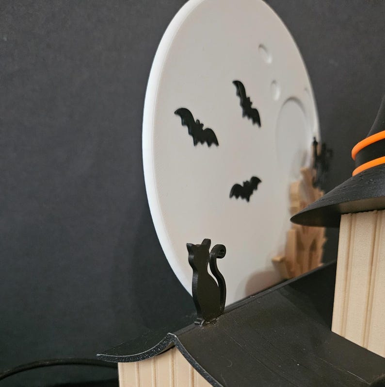 May include: A Halloween-themed decoration featuring a black cat on a rooftop, a witch's hat, and a white moon with black bat silhouettes. The scene is set against a dark background, creating a spooky ambiance.