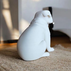 K&ouml;nnte beinhalten: Eine wei&szlig;e Bulldoggen-Skulptur aus Keramik, sitzend auf einem strukturierten, hellbraunen Teppich. Die Hundefigur ist glatt und gl&auml;nzend, mit sichtbaren Details der Rasse. Die Skulptur ist ein dekoratives Wohnaccessoire.