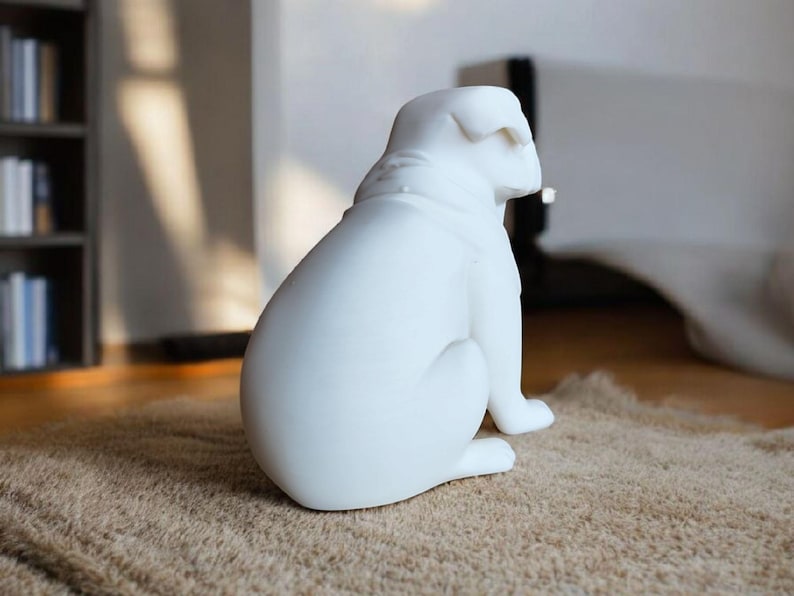 K&ouml;nnte beinhalten: Eine wei&szlig;e Bulldoggen-Skulptur aus Keramik, sitzend auf einem strukturierten, hellbraunen Teppich. Die Hundefigur ist glatt und gl&auml;nzend, mit sichtbaren Details der Rasse. Die Skulptur ist ein dekoratives Wohnaccessoire.