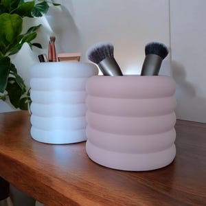 May include: Two cylindrical makeup brush holders. One is white, the other is pink. Both have a ribbed design and are filled with makeup brushes and small cosmetic products. The holders sit on a wooden surface.