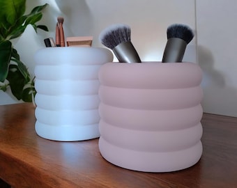 May include: Two cylindrical makeup brush holders. One is white, the other is pink. Both have a ribbed design and are filled with makeup brushes and small cosmetic products. The holders sit on a wooden surface.