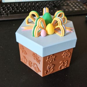 May include: A decorative hexagonal box with a light blue lid and brown base. The base features embossed floral and bunny designs. The lid is topped with colorful bow-shaped decorations and egg-shaped ornaments in green, yellow, and pink.