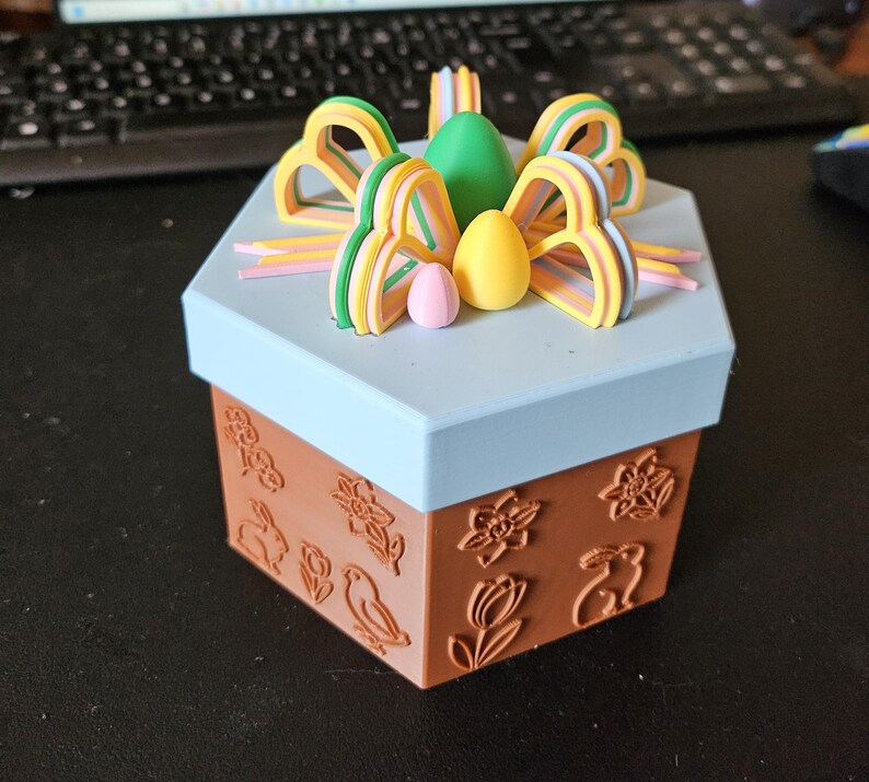 May include: A decorative hexagonal box with a light blue lid and brown base. The base features embossed floral and bunny designs. The lid is topped with colorful bow-shaped decorations and egg-shaped ornaments in green, yellow, and pink.