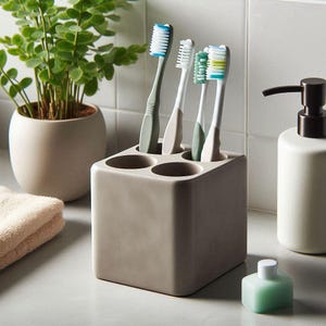 May include: A square, taupe toothbrush holder with four circular openings, holding four toothbrushes with blue, green, and white bristles. A white soap dispenser and a small green bottle are also visible. A potted plant and a folded towel are in the background.