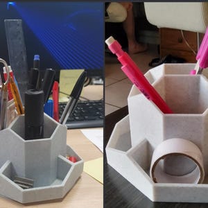 May include: A light gray desk organizer with multiple compartments for pens, pencils, and other office supplies. The organizer has a hexagonal shape with several angled storage areas. A roll of tape and various writing utensils are visible.