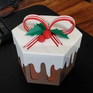 May include: A decorative, hexagonal box designed to resemble a Christmas pudding. The box features a white top with a brown base, and is adorned with a red and white striped bow, green holly leaves, and red berries. The box is a festive holiday decoration.