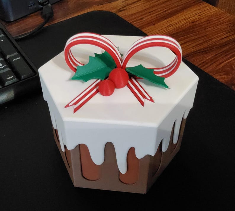 May include: A decorative, hexagonal box designed to resemble a Christmas pudding. The box features a white top with a brown base, and is adorned with a red and white striped bow, green holly leaves, and red berries. The box is a festive holiday decoration.