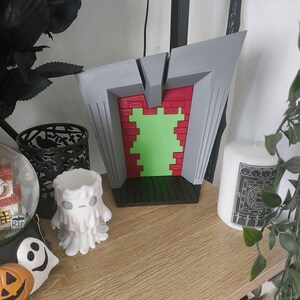 May include: A gray, red, and green decorative piece with a geometric design. The piece has a brick-like pattern in red and a green center. The design is set on a black base. A white candle and other Halloween decorations are nearby.