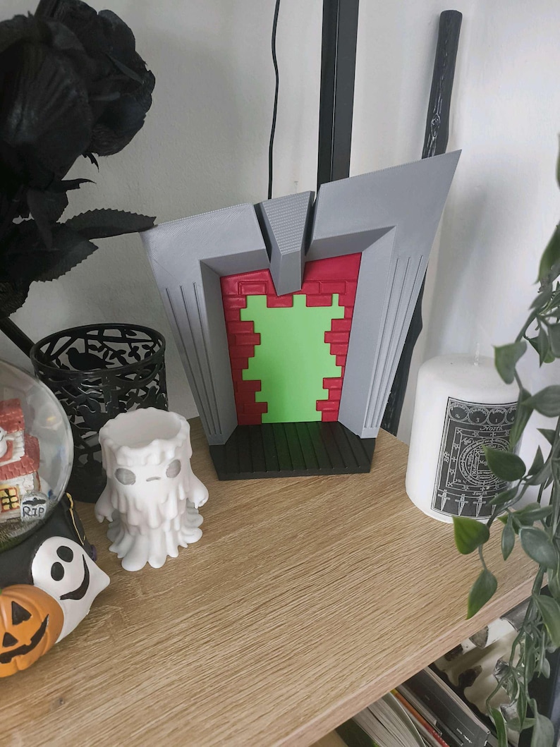 May include: A gray, red, and green decorative piece with a geometric design. The piece has a brick-like pattern in red and a green center. The design is set on a black base. A white candle and other Halloween decorations are nearby.