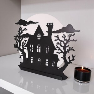 May include: Black metal haunted house silhouette with a white moon backdrop, trees, and clouds. A lit black candle sits to the right. The house has multiple windows and a chimney, creating a spooky Halloween decoration.