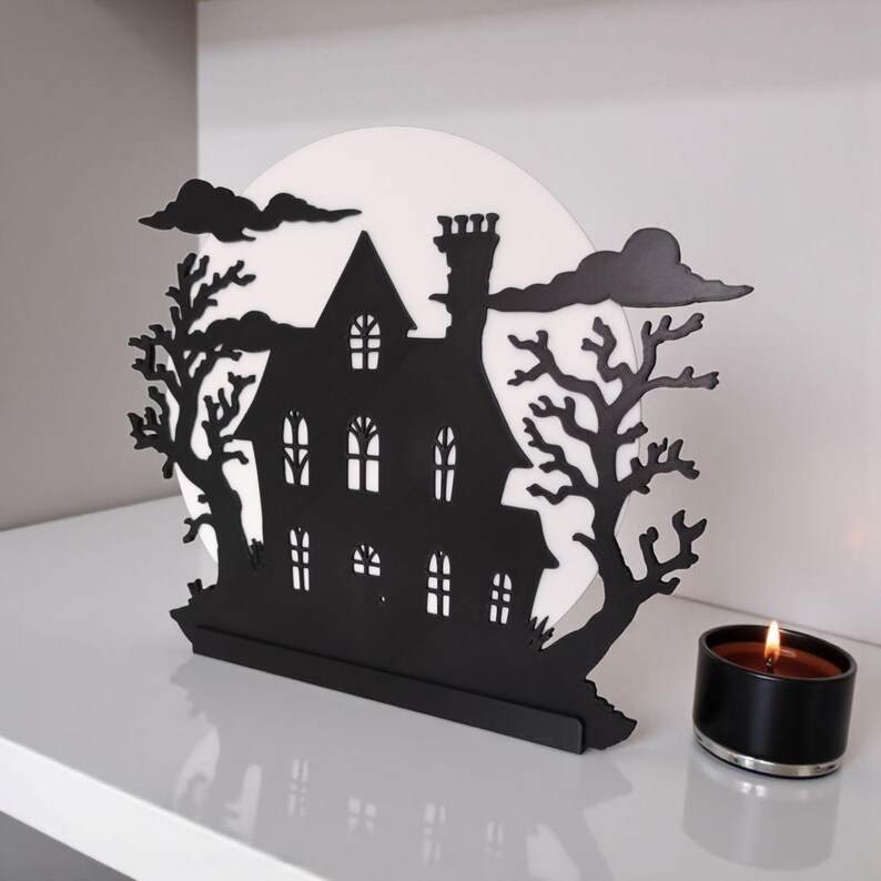 May include: Black metal haunted house silhouette with a white moon backdrop, trees, and clouds. A lit black candle sits to the right. The house has multiple windows and a chimney, creating a spooky Halloween decoration.