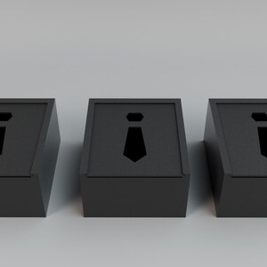 May include: Three black boxes with a tie-shaped cutout on the front. The boxes are angled and have a simple, modern design. The tie cutout is a solid black shape. The boxes are likely for storing small items.