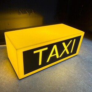 May include: A rectangular yellow taxi sign with the word "TAXI" in bold, illuminated letters. The sign has a black background for the text and is lit from within, creating a bright, eye-catching display. The sign is designed to be placed on a vehicle.