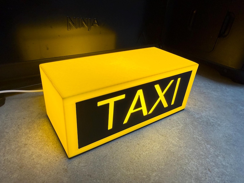 May include: A rectangular yellow taxi sign with the word "TAXI" in bold, illuminated letters. The sign has a black background for the text and is lit from within, creating a bright, eye-catching display. The sign is designed to be placed on a vehicle.