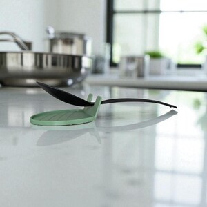 May include: A mint green spoon rest with a black spoon resting on it. The spoon rest has a circular base with a ridged design and a small handle to hold the spoon. The background shows a stainless steel pot and a window.