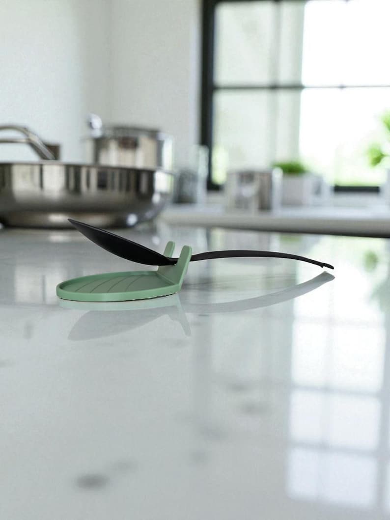 May include: A mint green spoon rest with a black spoon resting on it. The spoon rest has a circular base with a ridged design and a small handle to hold the spoon. The background shows a stainless steel pot and a window.