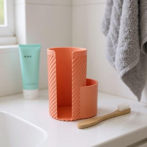 May include: A coral-colored toothbrush holder with a textured, diagonal stripe design. A small, round compartment is attached to the side. A wooden toothbrush with white bristles rests on the counter. A tube of teal-colored lotion is visible.