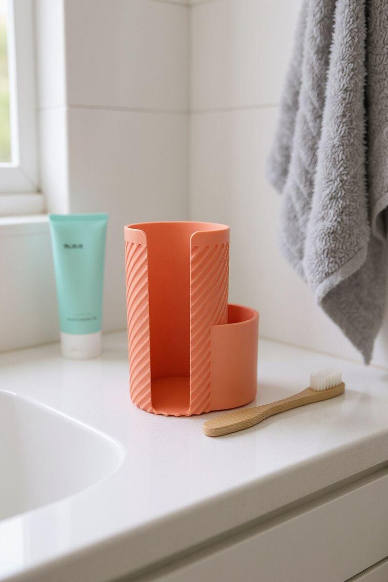 May include: A coral-colored toothbrush holder with a textured, diagonal stripe design. A small, round compartment is attached to the side. A wooden toothbrush with white bristles rests on the counter. A tube of teal-colored lotion is visible.