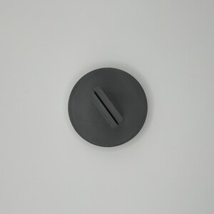 May include: A dark gray, circular object with a rectangular slot. The object appears to be a stand or holder, possibly for a phone or other small item. The design is simple and modern, with a matte finish.