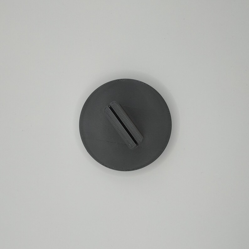 May include: A dark gray, circular object with a rectangular slot. The object appears to be a stand or holder, possibly for a phone or other small item. The design is simple and modern, with a matte finish.