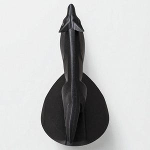 May include: A black decorative wall sculpture in the shape of a stylized animal, possibly a wolf or dog, viewed from above. The sculpture has a sleek, modern design and is mounted on a teardrop-shaped base. The artwork is made of a matte black material.