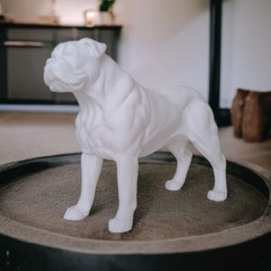 May include: A white bulldog statue, standing on a round, dark-colored surface. The sculpture is detailed, showing the dog's wrinkles and muscular build. The statue is a decorative item, likely for home decor.