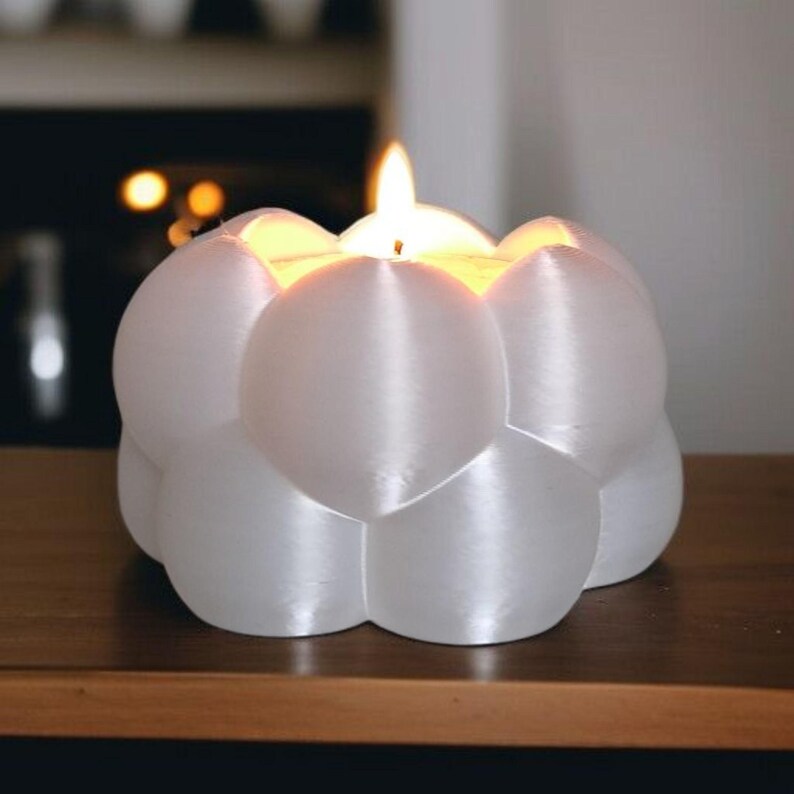 May include: A white, spherical candle holder with a lit candle. The holder has a bubbly, organic shape, and the flame glows brightly. The candle holder is on a wooden surface.