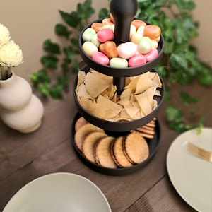 May include: A three-tiered black serving tray filled with snacks. The top tier holds colorful candies, the middle tier has potato chips, and the bottom tier contains cookies. The tray sits on a wooden surface, with plates and a vase in the background.