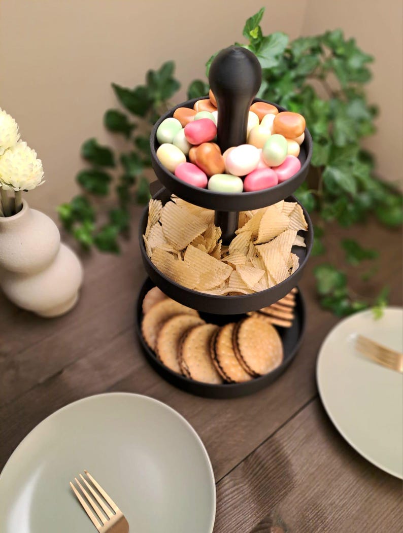 May include: A three-tiered black serving tray filled with snacks. The top tier holds colorful candies, the middle tier has potato chips, and the bottom tier contains cookies. The tray sits on a wooden surface, with plates and a vase in the background.