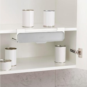 May include: A white cabinet interior with a pull-out storage shelf and several white cans. The shelf is gray and white, designed to hold items. The cabinet door is open, revealing the contents.