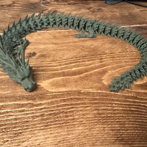May include: A 3D-printed articulated dragon toy in a muted green colour. The dragon is posed on a wooden surface, with its head and tail curved. The dragon has detailed scales and spikes along its back, and is a unique decorative item.
