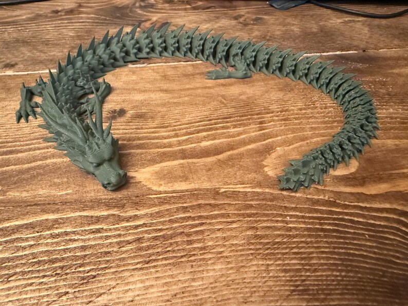 May include: A 3D-printed articulated dragon toy in a muted green colour. The dragon is posed on a wooden surface, with its head and tail curved. The dragon has detailed scales and spikes along its back, and is a unique decorative item.