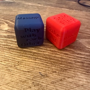 Puede incluir: Dos dados impresos en 3D, uno azul y otro rojo. El dado azul tiene las palabras "Massage" y "Play with each Other". El dado rojo tiene las palabras "Solo Play", "Wake Out" y "Massage".