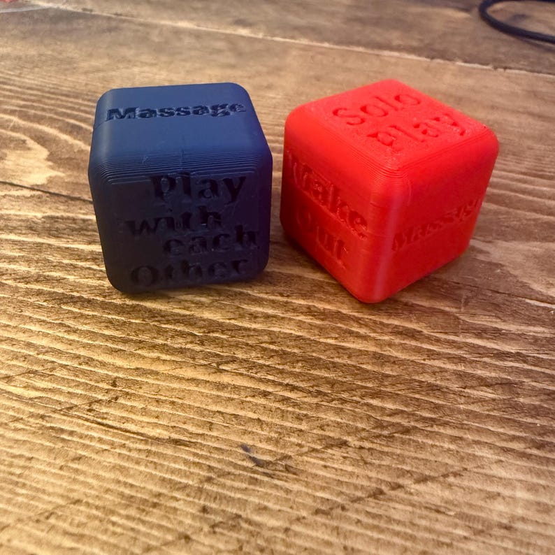 Puede incluir: Dos dados impresos en 3D, uno azul y otro rojo. El dado azul tiene las palabras "Massage" y "Play with each Other". El dado rojo tiene las palabras "Solo Play", "Wake Out" y "Massage".