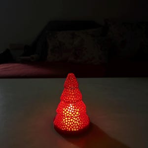 May include: A red, Christmas tree-shaped candle holder with a textured surface. The candle holder is lit from within, casting a warm glow and creating a long shadow on the surface.