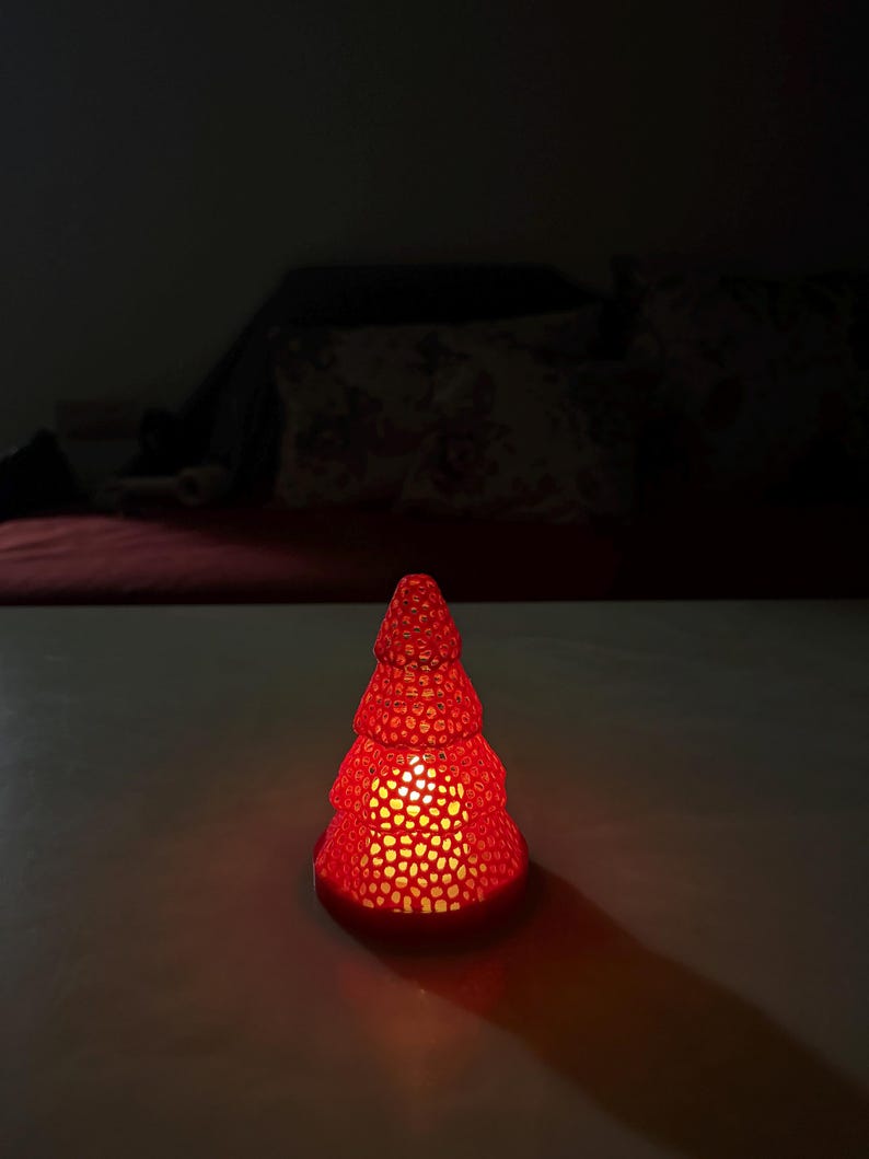May include: A red, Christmas tree-shaped candle holder with a textured surface. The candle holder is lit from within, casting a warm glow and creating a long shadow on the surface.