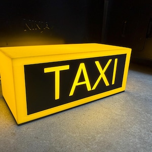May include: A rectangular yellow taxi sign with the word "TAXI" in bold, illuminated letters. The sign has a black background for the text and is lit from within, creating a bright, eye-catching display. The sign is on a gray surface.