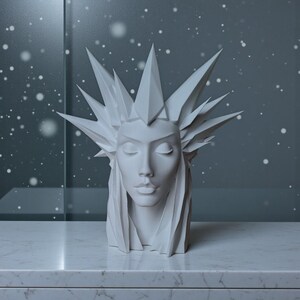 May include: A white sculpture of a serene face with closed eyes, adorned with a crown of sharp, angular spikes. The sculpture has long, flowing hair and is set against a blurred, snowy background. The sculpture is on a white marble surface.